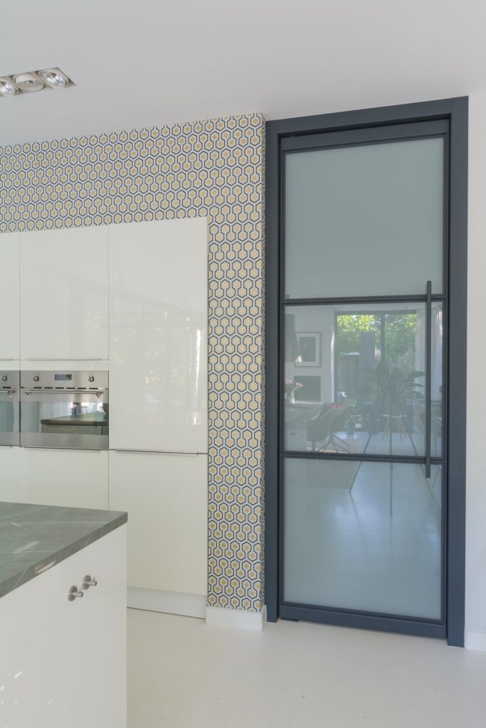 Stalen scharnierdeur met kozijnbekleding en melkglas toegepast in een moderne leefruimte met doorkijk naar de hal.