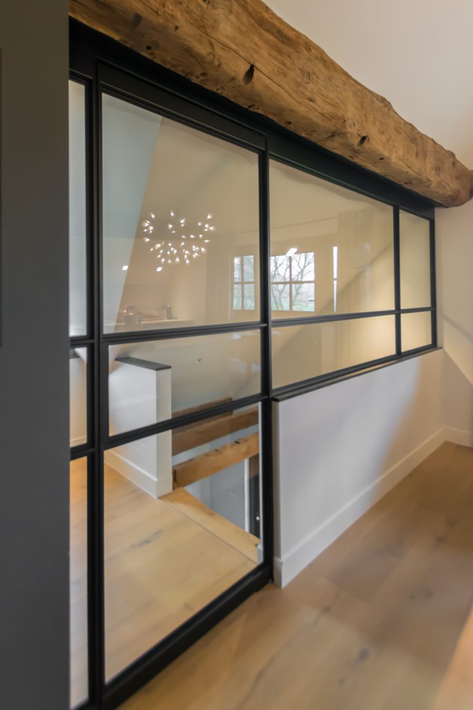 Stalen taatsdeur met slank zwart profiel onder een robuuste houten balk, met een moderne en lichte uitstraling.