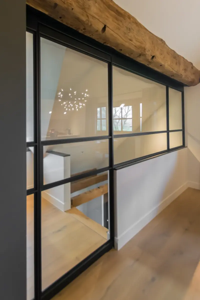 Stalen taatsdeur met raampaneel. Slank zwart profiel onder een robuuste houten balk, met een moderne en lichte uitstraling.
