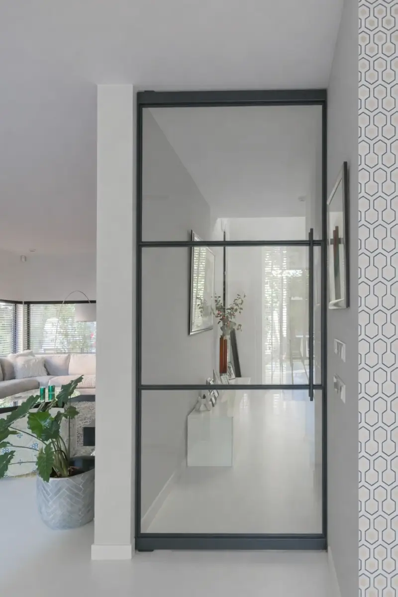 Stalen taatsdeur met slank zwart profiel die hal en woonkamer verbindt, met veel licht en een moderne, open uitstraling.